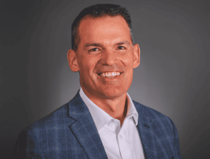 Portrait of a smiling middle-aged man in a blue blazer and white shirt against a gray background.