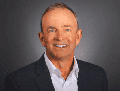 Headshot of a smiling middle-aged man with closely cropped grey hair, wearing a dark blazer and white shirt against a grey background.