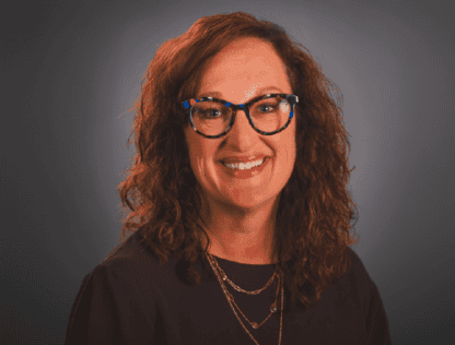 A middle-aged woman with curly brown hair and glasses, smiling in a professional portrait with a dark background.