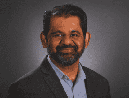 Portrait of a smiling middle-aged man with a beard, wearing a suit jacket and blue shirt, posed against a dark gray background.