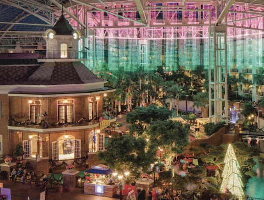 A bustling indoor atrium with lush tropical plants, a festive holiday tree adorned with lights, and a two-story building with a clock tower sets the stage for the Telarus Partner Summit 2024. The ceiling features a grid of colorful, illuminated patterns in green and pink, creating a vibrant, lively atmosphere.