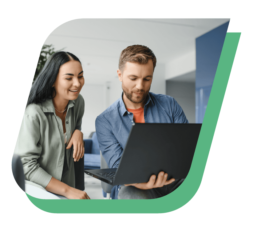 A man and woman sit together, looking at a laptop screen. The man, in a blue shirt, holds the laptop, while the woman, in a green shirt, smiles and leans in to look. They appear to be breaking barriers in a modern, brightly lit workspace, framed by a green curved border.