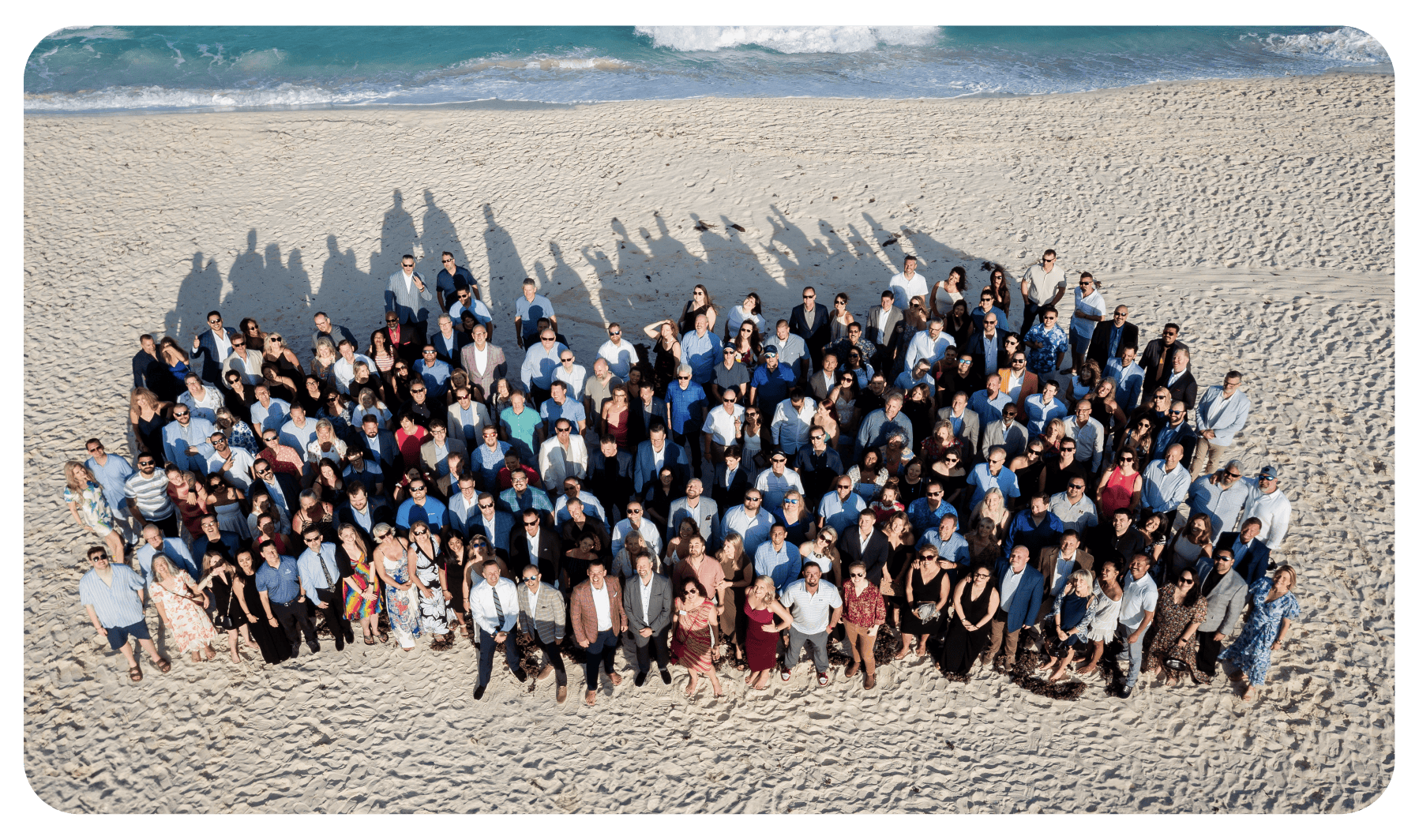 A large group of people, symbolizing the spirit of growth, stands closely together on a sandy beach, with the ocean visible in the background. They wear a mix of formal and casual outfits under clear skies, their shadows stretching out on the sand behind them.