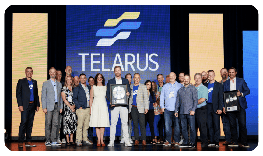 A group of people stands proudly on a stage with the Telarus logo displayed on a large screen behind them. Celebrating innovation, some individuals hold awards. The smiling team, dressed in business and casual attire, is framed by colorful vertical panels on either side.