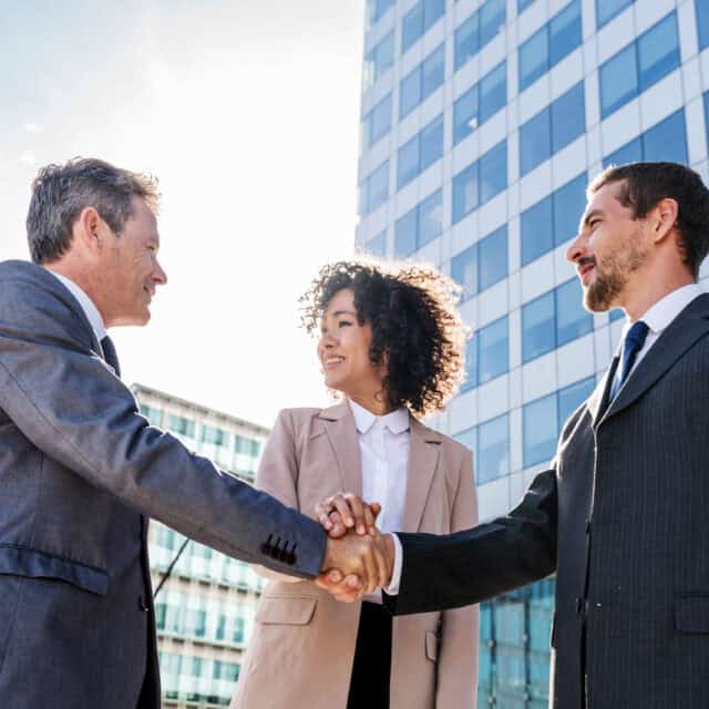 Standing outside a towering building, two business professionals in sleek suits are shaking hands with Telarus Capital's representative, a poised woman in a smart blazer, smiling warmly. The bright sky serves as their backdrop, casting a hopeful glow over this promising partnership.